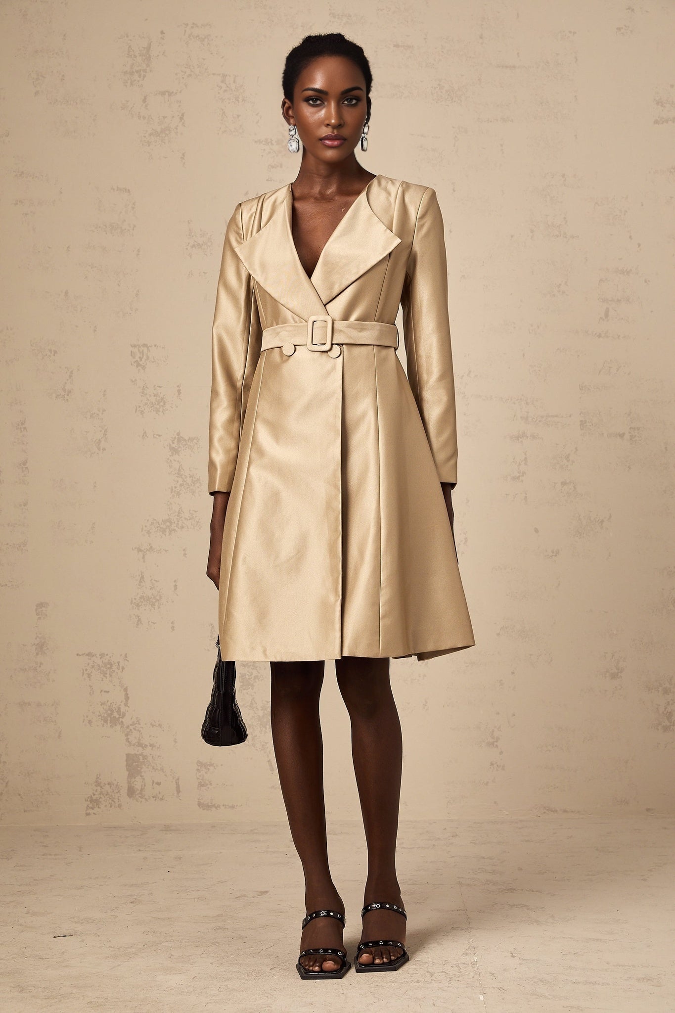 Woman wearing long khaki coat with belted waist and open front standing against neutral background