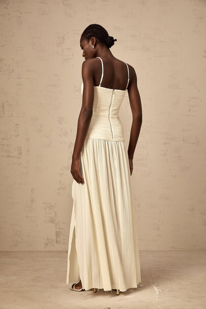Woman wearing long neutral colored pleated maxi dress with side slit standing in relaxed pose
