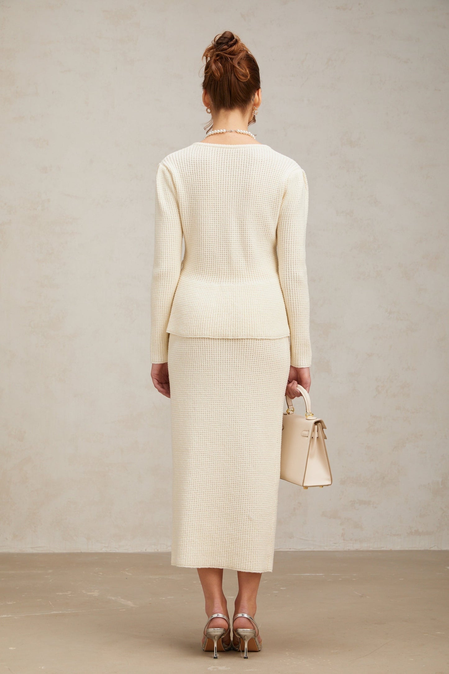 A woman wearing a white knitted matching set consisting of a sleeveless top and a knee length skirt standing against a plain background