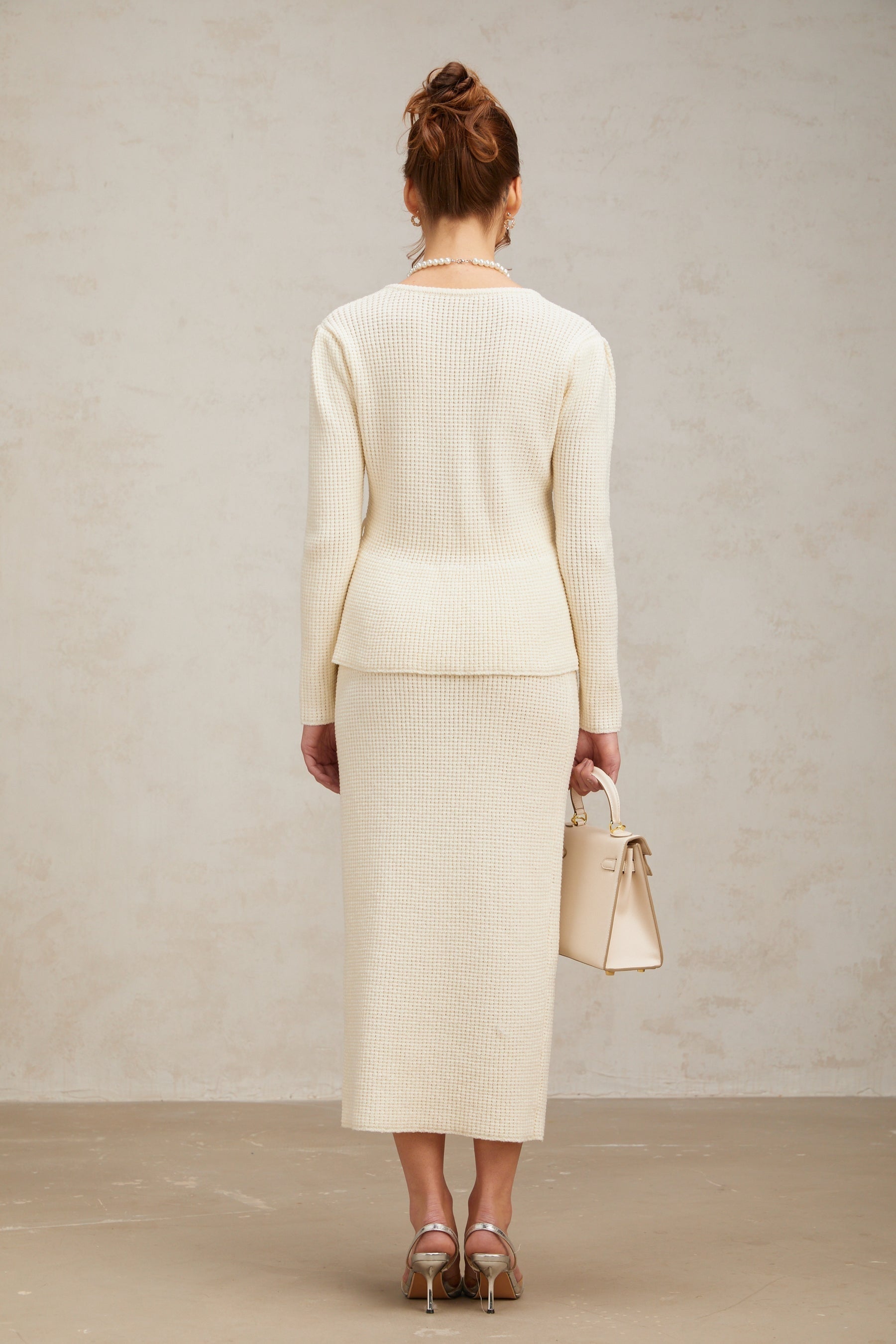 A woman wearing a white knitted matching set consisting of a sleeveless top and a knee length skirt standing against a plain background