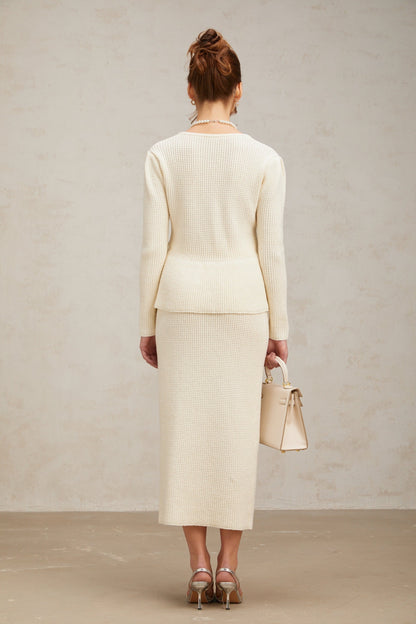 A woman wearing a white knitted matching set consisting of a sleeveless top and a knee length skirt standing against a plain background
