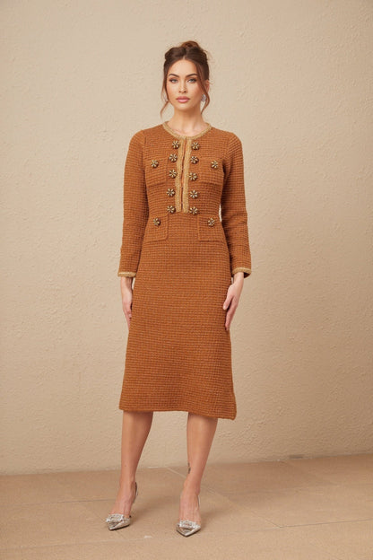 A woman wearing a brown tweed dress with braided edging standing against a plain background
