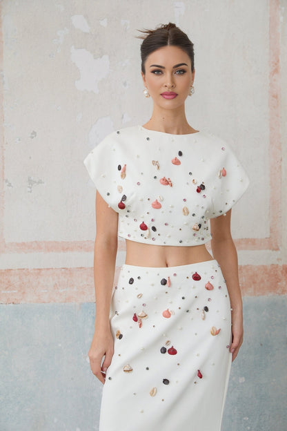 A white twopiece outfit consisting of a sleeveless top and skirt decorated with shell embellishments matching set called Delphine