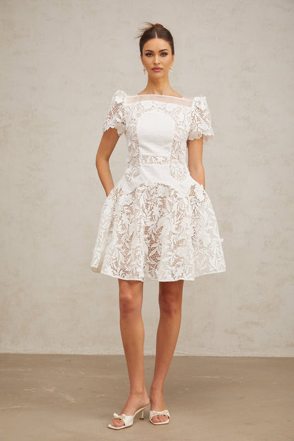 Young woman wearing a white mini dress with floral lace pattern and puff sleeves standing against a plain background
