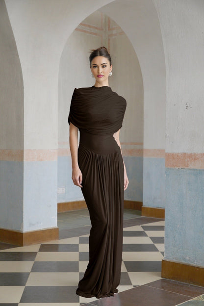 A brown asymmetric maxi dress with pleated design and flowing skirt worn by a female model standing against a plain background