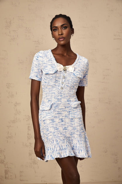A young woman wearing a blue and white checked knit mini dress with short sleeves and a fitted silhouette standing against a plain background
