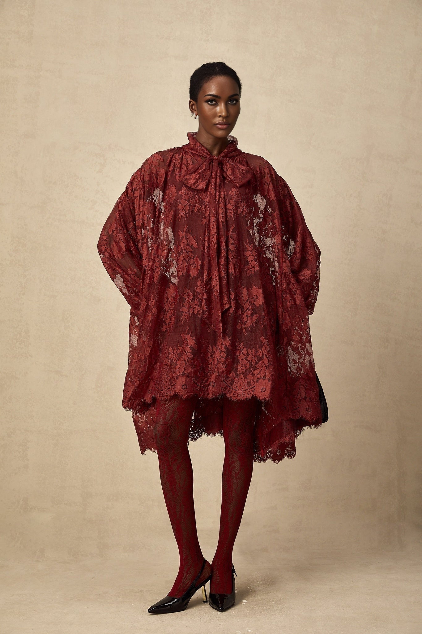 A woman wearing a flowing red oversize dress made of delicate chantilly lace standing against a plain background